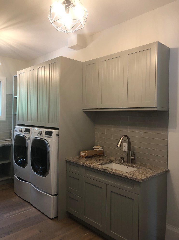 Foto på en vintage tvättstuga, med en undermonterad diskho, grå skåp, granitbänkskiva, beige väggar, mörkt trägolv, en tvättmaskin och torktumlare bredvid varandra och brunt golv