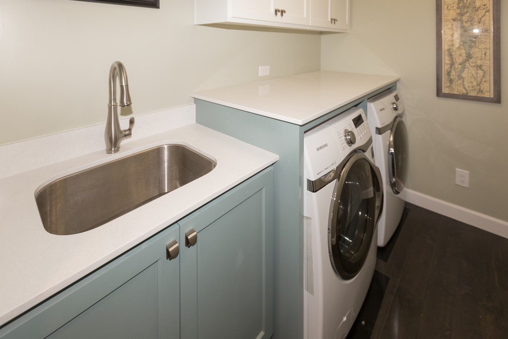 Klassisk inredning av en mellanstor parallell tvättstuga enbart för tvätt, med en undermonterad diskho, skåp i shakerstil, bänkskiva i kvarts, grå väggar, mörkt trägolv, en tvättmaskin och torktumlare bredvid varandra och blå skåp
