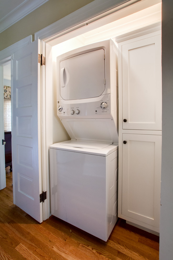 Kleiner Klassischer Hauswirtschaftsraum mit Waschmaschinenschrank, Schrankfronten im Shaker-Stil, Waschmaschine und Trockner gestapelt, weißer Wandfarbe und braunem Holzboden in Los Angeles