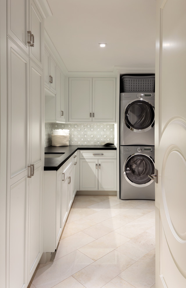 マイアミにあるトランジショナルスタイルのおしゃれな洗濯室 (L型、アンダーカウンターシンク、落し込みパネル扉のキャビネット、白いキャビネット、上下配置の洗濯機・乾燥機、ベージュの床、黒いキッチンカウンター) の写真