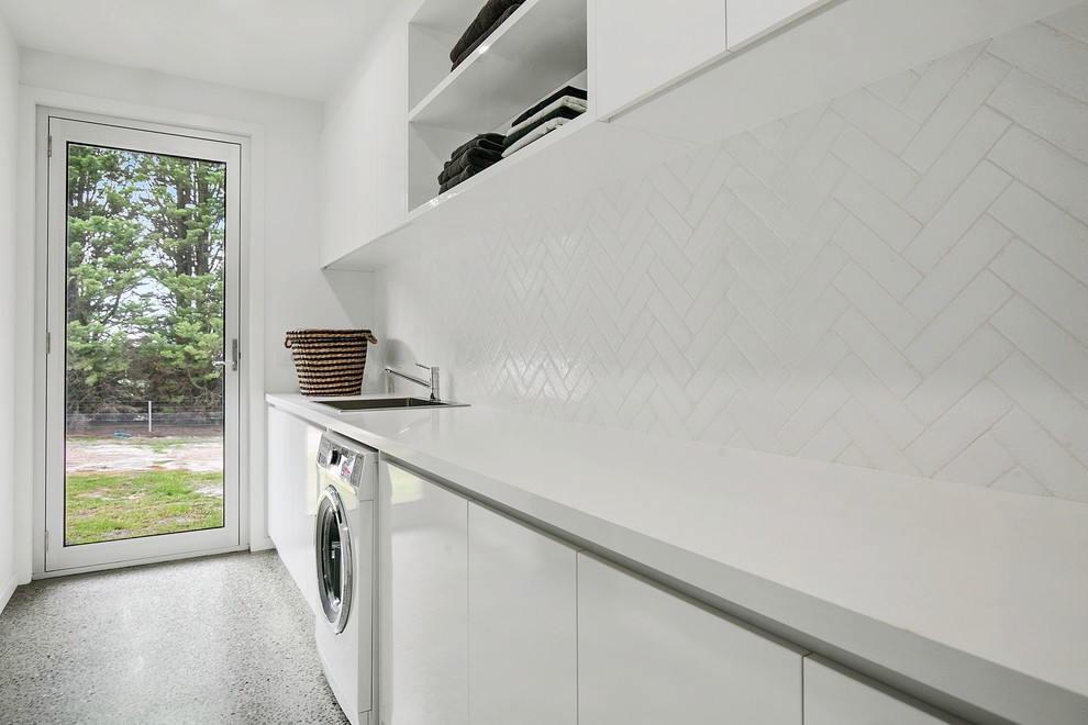 Inspiration för en mycket stor funkis linjär tvättstuga enbart för tvätt, med en nedsänkt diskho, släta luckor, vita skåp, bänkskiva i kvarts, vita väggar och betonggolv