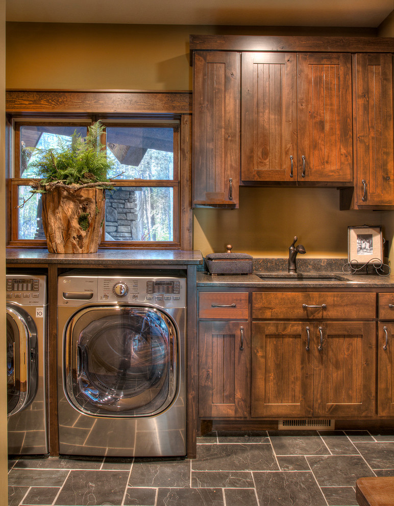Bild på en mellanstor rustik grå linjär grått tvättstuga enbart för tvätt, med en undermonterad diskho, skåp i mellenmörkt trä, granitbänkskiva, skiffergolv, en tvättmaskin och torktumlare bredvid varandra, grått golv, skåp i shakerstil och bruna väggar