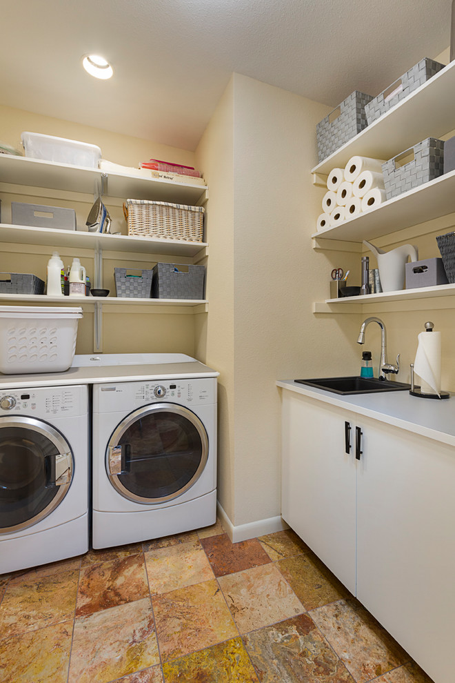 Bild på en liten funkis l-formad tvättstuga enbart för tvätt, med en nedsänkt diskho, öppna hyllor, vita skåp, laminatbänkskiva, beige väggar, travertin golv, en tvättmaskin och torktumlare bredvid varandra och flerfärgat golv