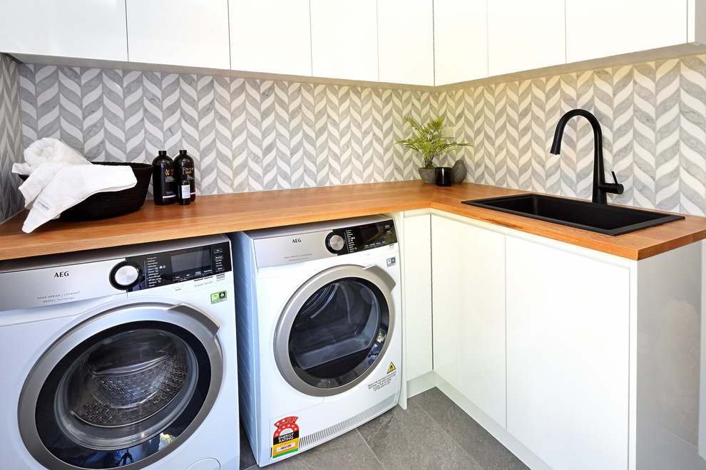 Exempel på en liten modern bruna l-formad brunt tvättstuga enbart för tvätt, med en undermonterad diskho, luckor med glaspanel, vita skåp, träbänkskiva, grå väggar, klinkergolv i keramik, en tvättmaskin och torktumlare bredvid varandra och grått golv