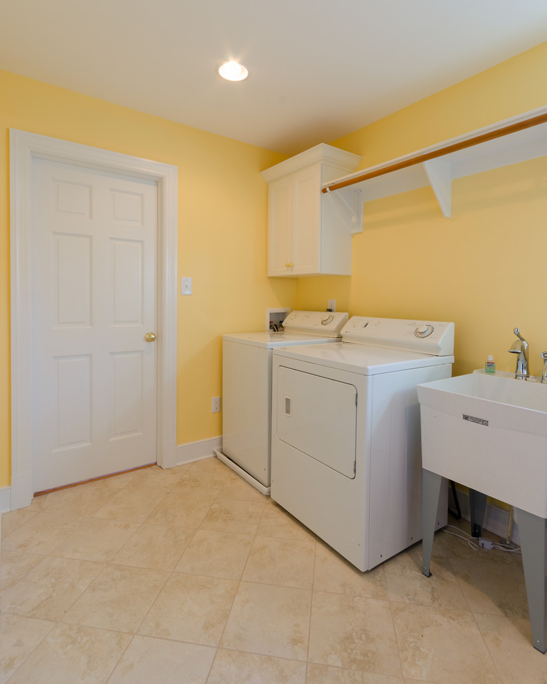 Modelo de cuarto de lavado lineal tradicional de tamaño medio con pila para lavar, paredes amarillas, suelo de baldosas de cerámica, lavadora y secadora juntas y suelo beige
