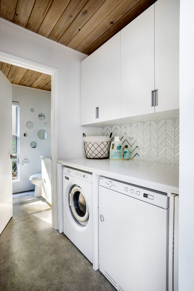 Foto på en liten retro vita linjär tvättstuga enbart för tvätt, med släta luckor, vita skåp, bänkskiva i kvarts, vita väggar, betonggolv, tvättmaskin och torktumlare byggt in i ett skåp och grått golv