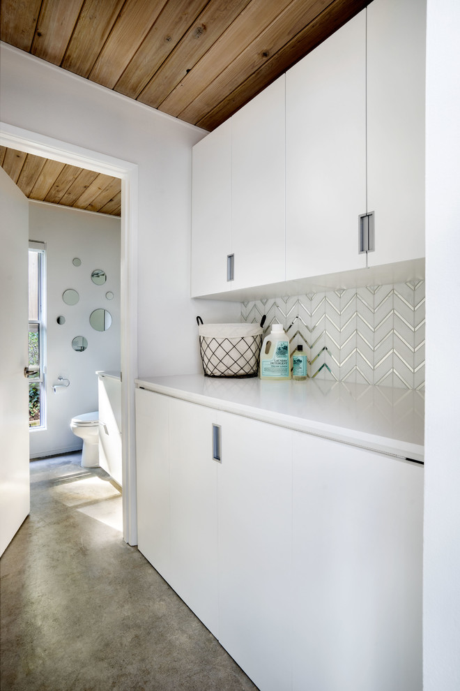 Small 1950s single-wall concrete floor laundry room photo in Portland with flat-panel cabinets, white cabinets, quartz countertops, white walls and a concealed washer/dryer