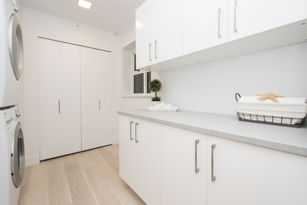 Example of a mid-sized trendy galley light wood floor and beige floor dedicated laundry room design in Vancouver with flat-panel cabinets, white cabinets, quartz countertops, white walls, a stacked washer/dryer and gray countertops