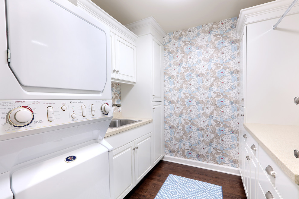 Design ideas for a medium sized classic galley laundry cupboard in Grand Rapids with a single-bowl sink, raised-panel cabinets, white cabinets, engineered stone countertops, multi-coloured walls, dark hardwood flooring and a stacked washer and dryer.