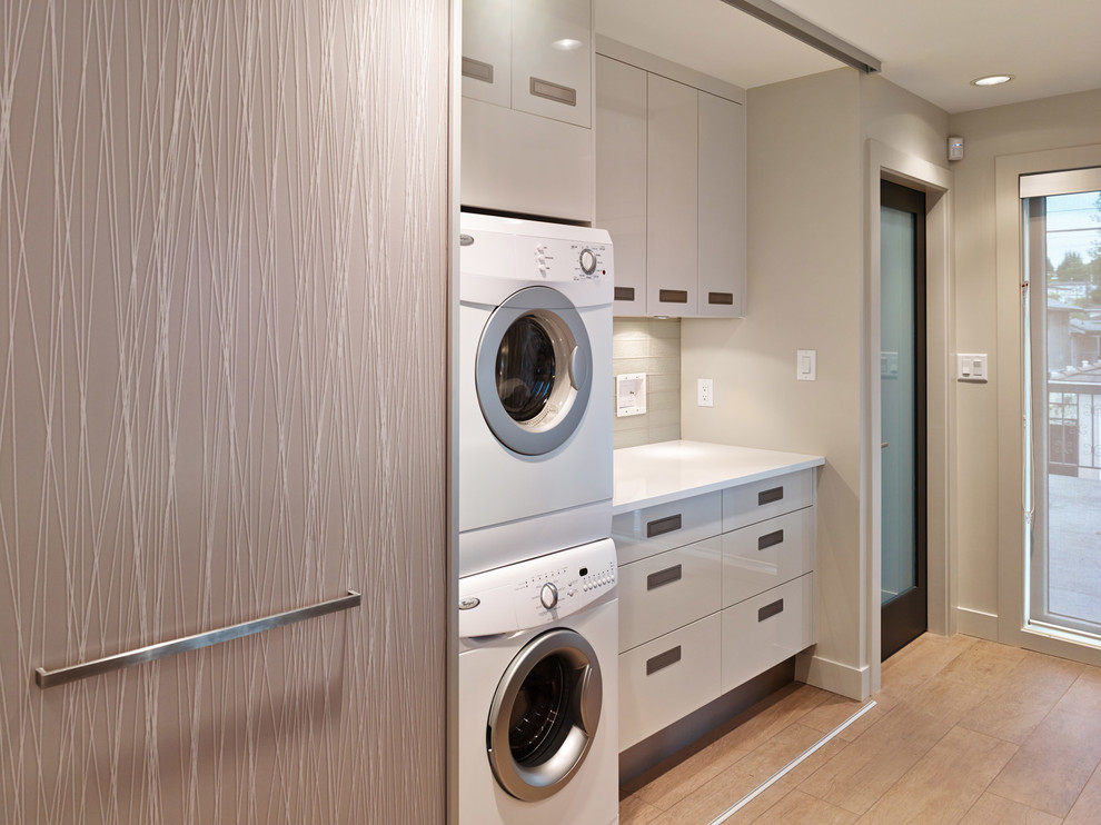 Modelo de lavadero contemporáneo con armarios con paneles lisos, puertas de armario grises, paredes beige, suelo de madera en tonos medios, lavadora y secadora apiladas y encimeras blancas
