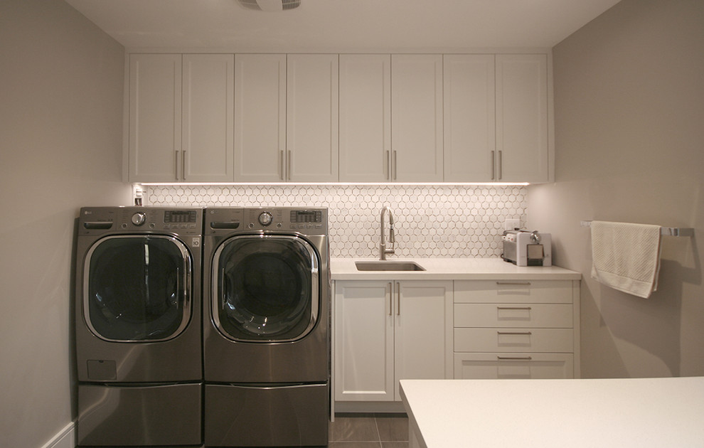 Inredning av en modern stor vita parallell vitt tvättstuga enbart för tvätt, med en undermonterad diskho, skåp i shakerstil, vita skåp, bänkskiva i kvartsit, beige väggar, betonggolv, en tvättmaskin och torktumlare bredvid varandra och grått golv