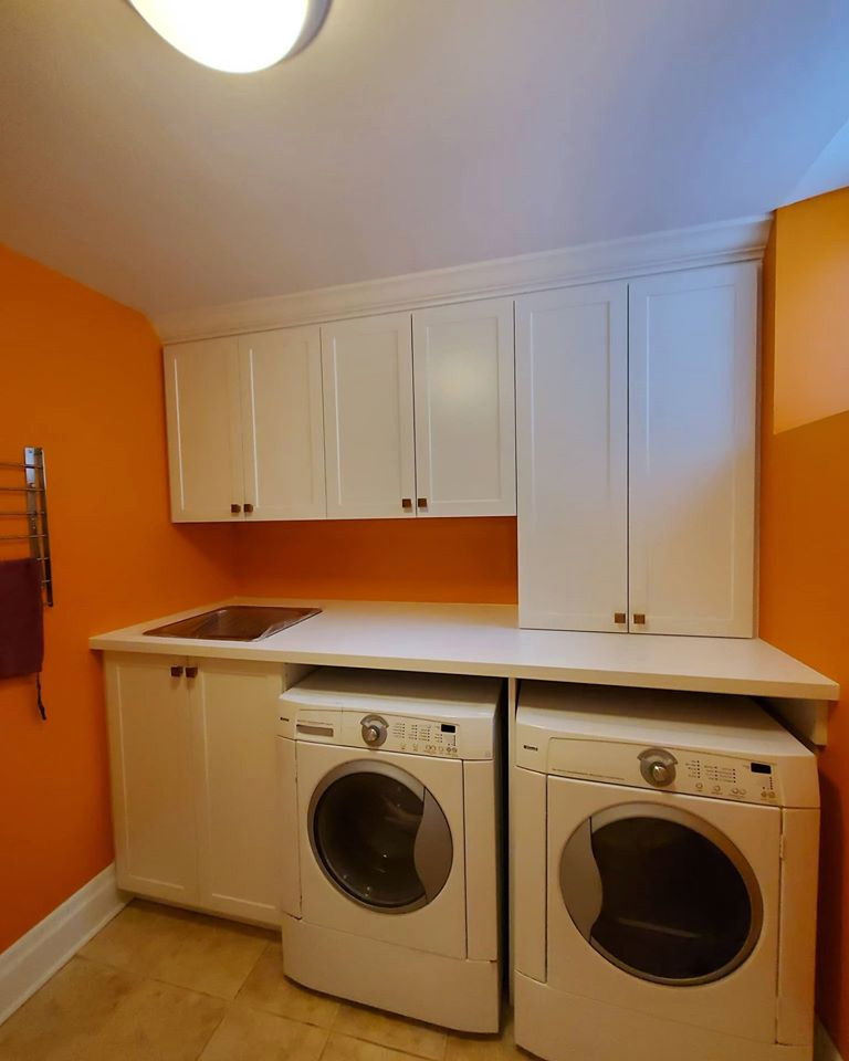 Inredning av en vita linjär vitt tvättstuga, med skåp i shakerstil, vita skåp, orange väggar och en tvättmaskin och torktumlare bredvid varandra