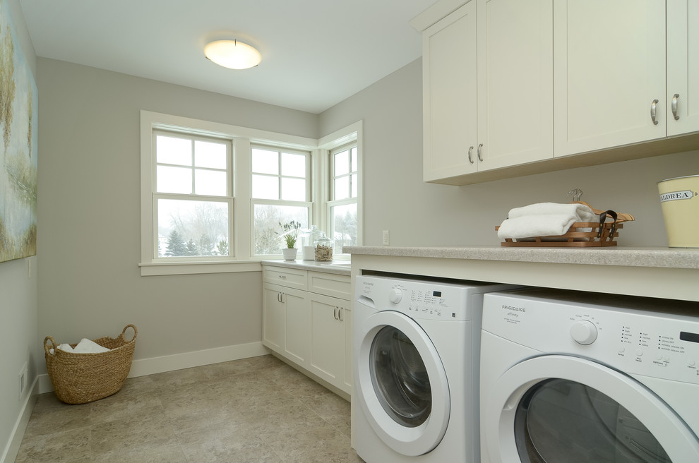 Modelo de cuarto de lavado lineal de estilo de casa de campo con armarios estilo shaker, puertas de armario blancas, paredes grises y lavadora y secadora juntas