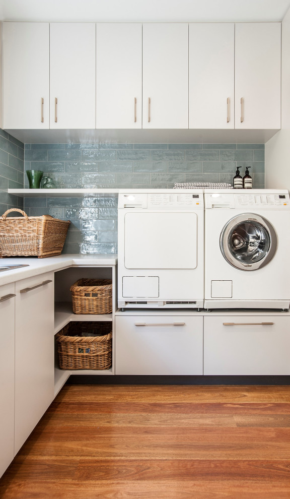 Modern inredning av en stor grå u-formad grått tvättstuga enbart för tvätt, med en dubbel diskho, vita skåp, laminatbänkskiva, blått stänkskydd, stänkskydd i tunnelbanekakel, vita väggar, mellanmörkt trägolv, en tvättmaskin och torktumlare bredvid varandra och brunt golv