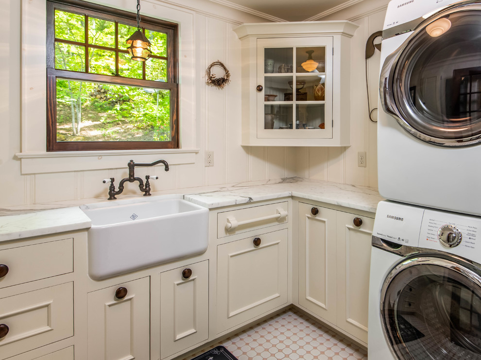 Photo of a small rural l-shaped separated utility room in Other with a belfast sink, recessed-panel cabinets, beige cabinets, marble worktops, beige walls, ceramic flooring, a stacked washer and dryer and pink floors.