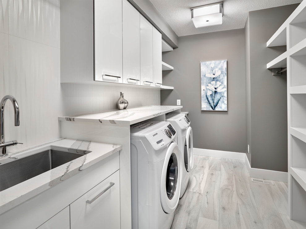 Große Moderne Waschküche mit Unterbauwaschbecken, Lamellenschränken, weißen Schränken, Quarzwerkstein-Arbeitsplatte, grauer Wandfarbe, Porzellan-Bodenfliesen, Waschmaschine und Trockner nebeneinander, grauem Boden und weißer Arbeitsplatte in Edmonton