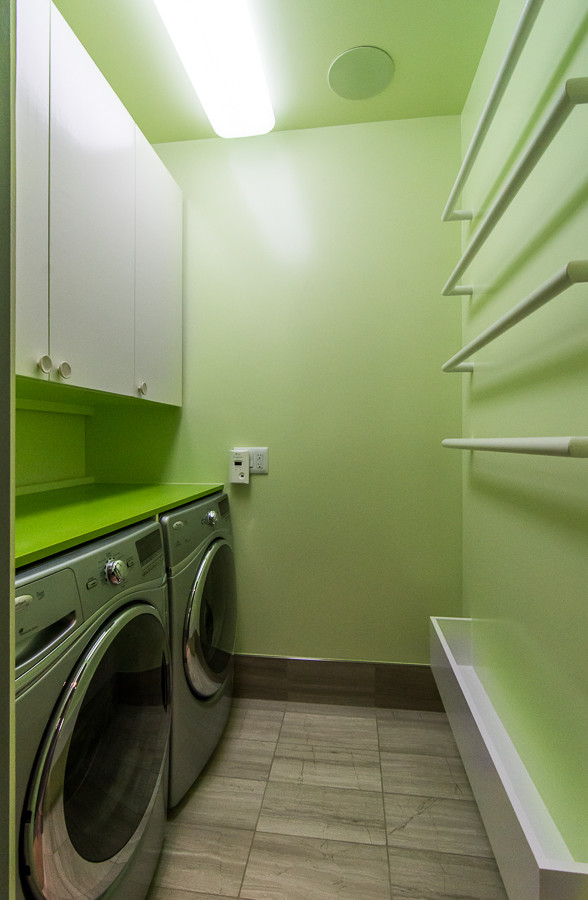 Inredning av en modern mellanstor gröna linjär grönt tvättstuga enbart för tvätt, med släta luckor, vita skåp, gröna väggar, klinkergolv i keramik, en tvättmaskin och torktumlare bredvid varandra och grått golv