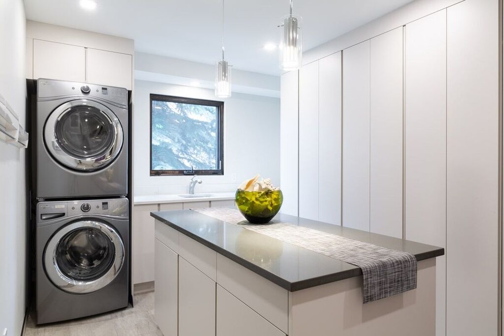 Ejemplo de cuarto de lavado actual con fregadero bajoencimera, armarios con paneles lisos, puertas de armario blancas, paredes blancas, lavadora y secadora apiladas y encimeras blancas