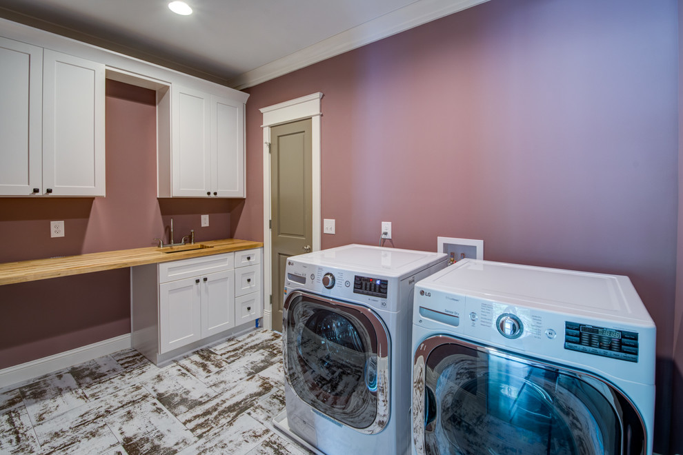 Inredning av en klassisk stor beige linjär beige tvättstuga enbart för tvätt, med en undermonterad diskho, skåp i shakerstil, vita skåp, träbänkskiva, en tvättmaskin och torktumlare bredvid varandra, vitt golv och lila väggar