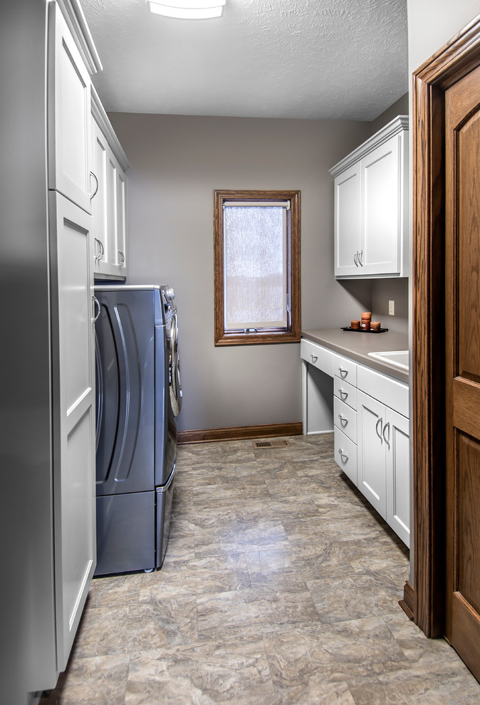 Modelo de cuarto de lavado de tamaño medio con armarios con paneles empotrados, puertas de armario blancas, encimera de laminado, paredes grises, suelo de linóleo y lavadora y secadora juntas
