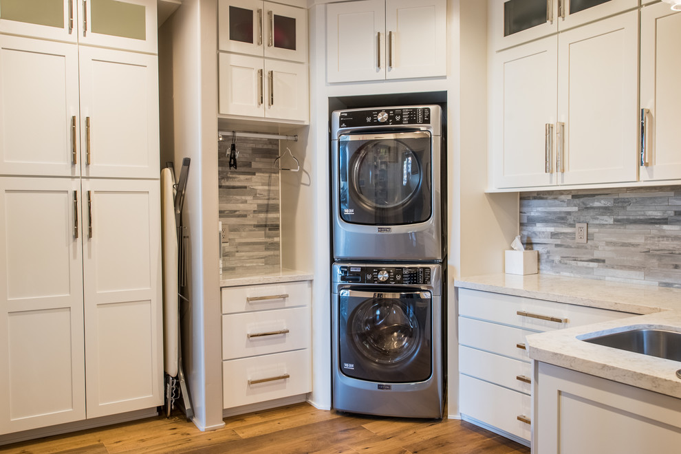 This is an example of a large rustic u-shaped utility room in Phoenix with a submerged sink, shaker cabinets, white cabinets, black walls, medium hardwood flooring, a stacked washer and dryer, brown floors and white worktops.