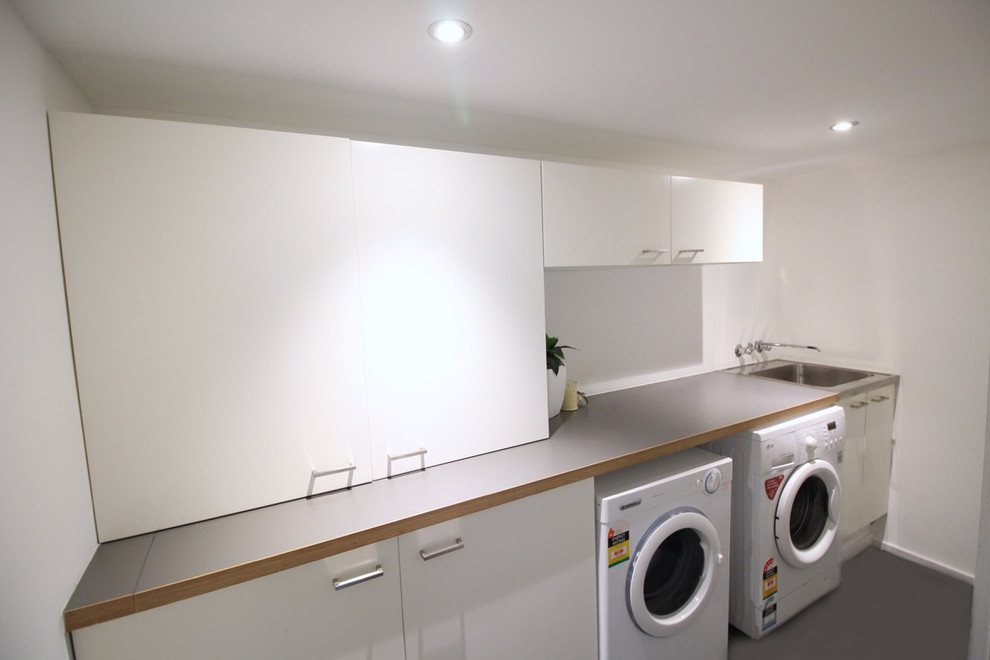 Modern inredning av en mellanstor parallell tvättstuga enbart för tvätt, med vita skåp, vita väggar, betonggolv och en tvättmaskin och torktumlare bredvid varandra