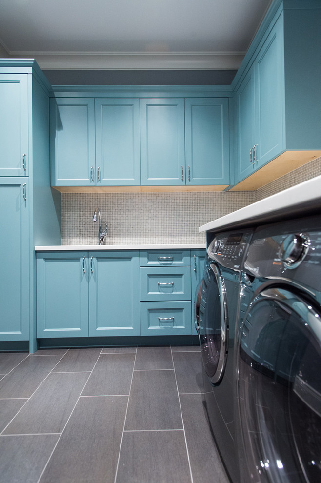 Klassisk inredning av en stor l-formad tvättstuga enbart för tvätt, med en undermonterad diskho, luckor med infälld panel, blå skåp, bänkskiva i kvarts, grå väggar, skiffergolv och en tvättmaskin och torktumlare bredvid varandra