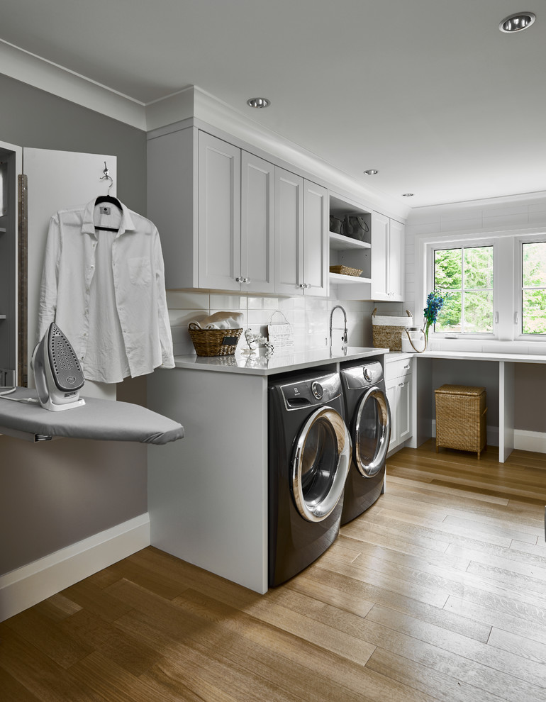 Idéer för en mellanstor klassisk vita l-formad tvättstuga enbart för tvätt, med skåp i shakerstil, bänkskiva i kvarts, grå väggar, ljust trägolv, grå skåp och en tvättmaskin och torktumlare bredvid varandra