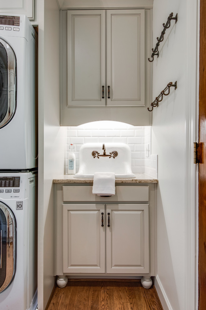 Photo of a large farmhouse galley utility room in Nashville with a single-bowl sink, recessed-panel cabinets, white cabinets, granite worktops, white walls, medium hardwood flooring, a stacked washer and dryer, brown floors and beige worktops.