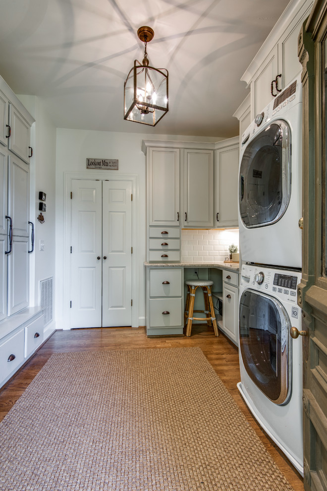 Utility room - large farmhouse galley medium tone wood floor and brown floor utility room idea in Nashville with a single-bowl sink, recessed-panel cabinets, white cabinets, granite countertops, white walls, a stacked washer/dryer and beige countertops