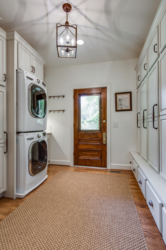 Utility room - large cottage galley medium tone wood floor and brown floor utility room idea in Nashville with recessed-panel cabinets, white cabinets, granite countertops, white walls, a stacked washer/dryer and beige countertops