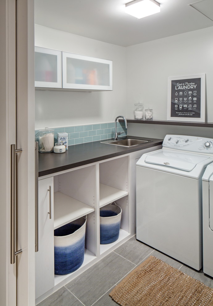 Foto på en liten funkis grå l-formad tvättstuga enbart för tvätt, med en nedsänkt diskho, öppna hyllor, vita skåp, laminatbänkskiva, vita väggar, klinkergolv i porslin och en tvättmaskin och torktumlare bredvid varandra