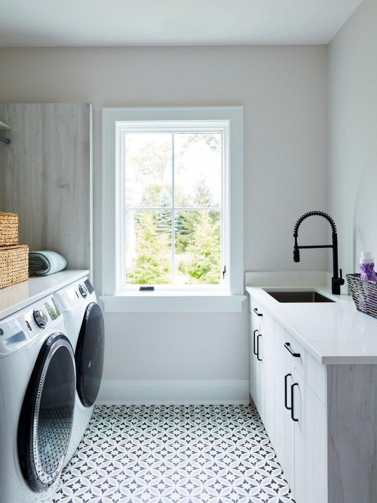 Klassisk inredning av en liten grå parallell grått tvättstuga enbart för tvätt, med en undermonterad diskho, släta luckor, grå skåp, bänkskiva i kvarts, grå väggar, klinkergolv i porslin, en tvättmaskin och torktumlare bredvid varandra och flerfärgat golv