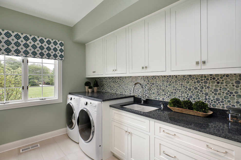 Inredning av en klassisk linjär tvättstuga enbart för tvätt, med en dubbel diskho, luckor med infälld panel, vita skåp, granitbänkskiva, klinkergolv i porslin, en tvättmaskin och torktumlare bredvid varandra och grå väggar