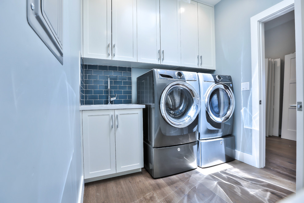 Foto på en mellanstor funkis grå parallell tvättstuga enbart för tvätt, med en undermonterad diskho, skåp i shakerstil, vita skåp, bänkskiva i kvarts, blå väggar, mellanmörkt trägolv och en tvättmaskin och torktumlare bredvid varandra