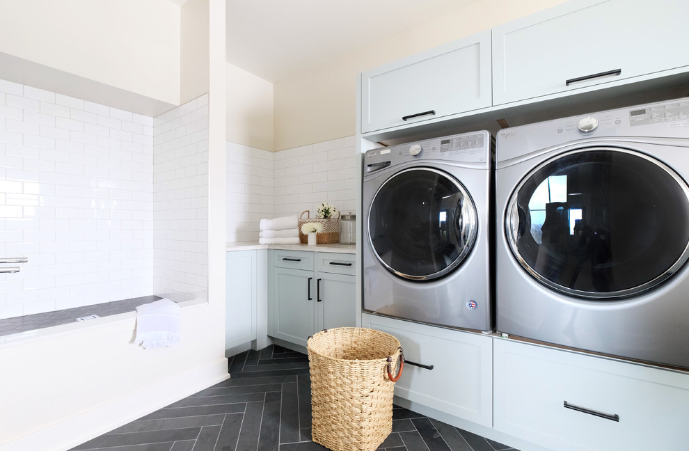 Modelo de cuarto de lavado lineal de estilo de casa de campo de tamaño medio con armarios estilo shaker, puertas de armario blancas, lavadora y secadora juntas, suelo gris y encimeras beige
