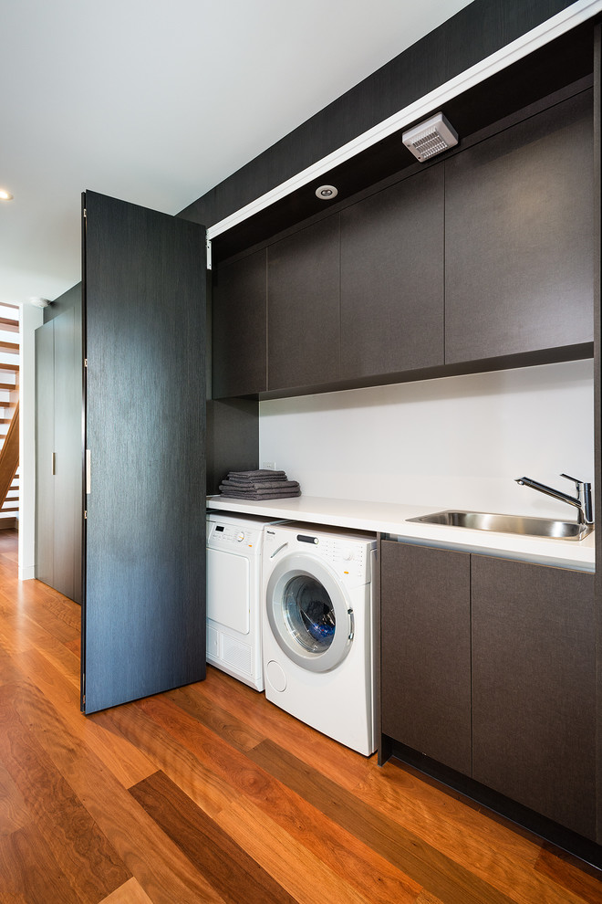 Einzeiliger, Mittelgroßer Moderner Hauswirtschaftsraum mit Waschmaschinenschrank, Einbauwaschbecken, Quarzwerkstein-Arbeitsplatte, weißer Wandfarbe, braunem Holzboden, Waschmaschine und Trockner nebeneinander, flächenbündigen Schrankfronten, weißer Arbeitsplatte und braunen Schränken in Melbourne