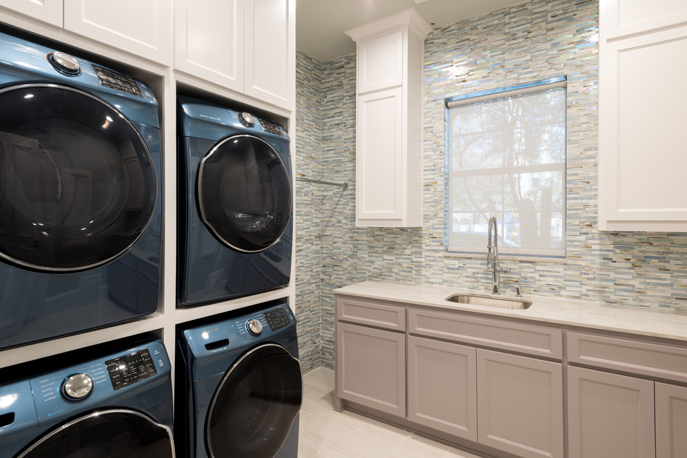 Large transitional galley porcelain tile and beige floor dedicated laundry room photo in Dallas with an undermount sink, recessed-panel cabinets, beige cabinets, multicolored walls, a stacked washer/dryer and marble countertops