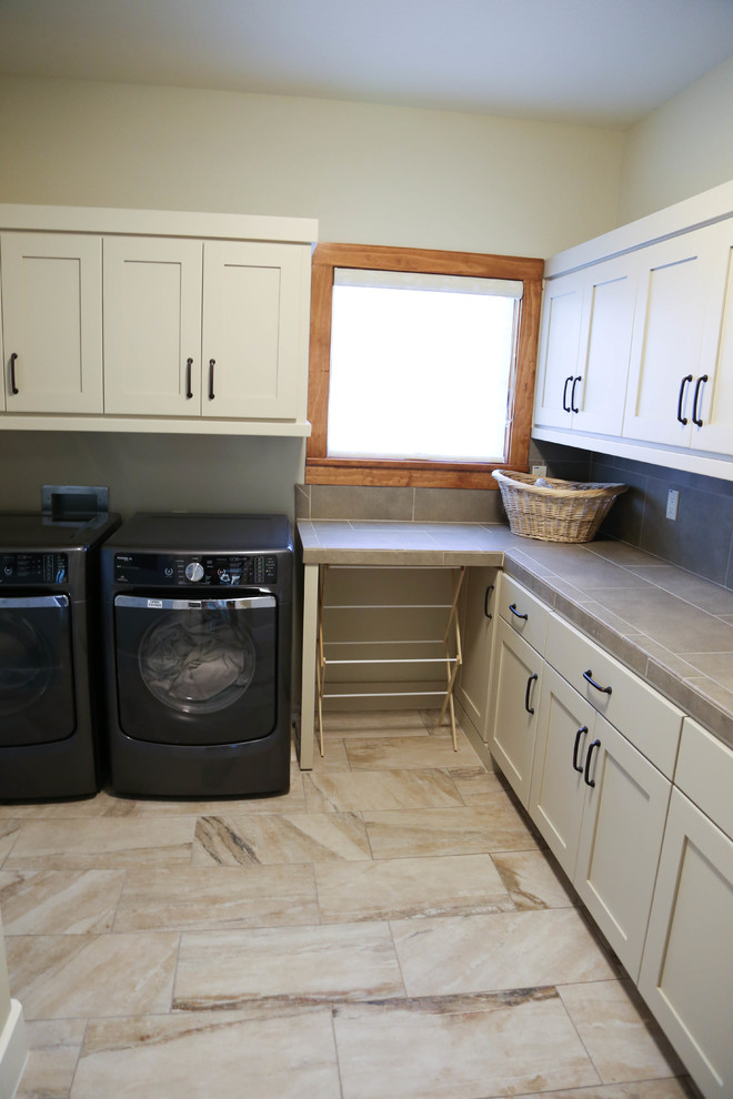Inspiration for a medium sized contemporary l-shaped separated utility room in Austin with a side by side washer and dryer, shaker cabinets, beige cabinets, tile countertops, beige walls and travertine flooring.