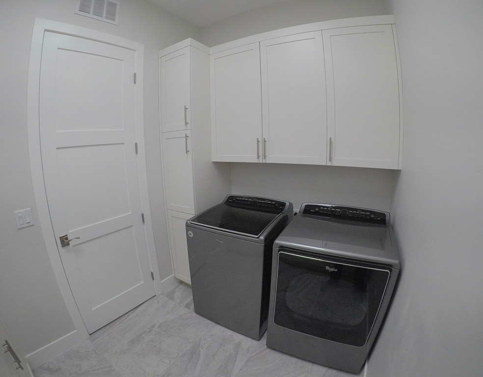 Modern inredning av en mellanstor parallell tvättstuga enbart för tvätt, med en nedsänkt diskho, skåp i shakerstil, vita skåp, granitbänkskiva, vita väggar, klinkergolv i porslin, en tvättmaskin och torktumlare bredvid varandra och grått golv