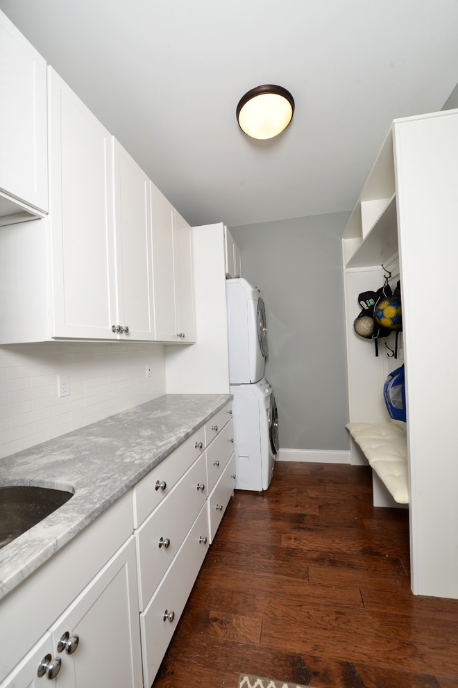 Small transitional single-wall medium tone wood floor and brown floor utility room photo in St Louis with an undermount sink, shaker cabinets, white cabinets, marble countertops, gray walls, a stacked washer/dryer and gray countertops