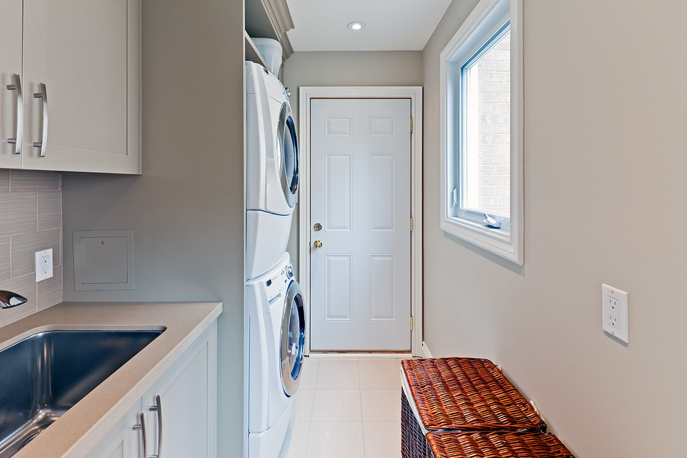 This is an example of a small galley utility room in Toronto with a submerged sink, shaker cabinets, grey cabinets, engineered stone countertops, grey walls, marble flooring and a stacked washer and dryer.