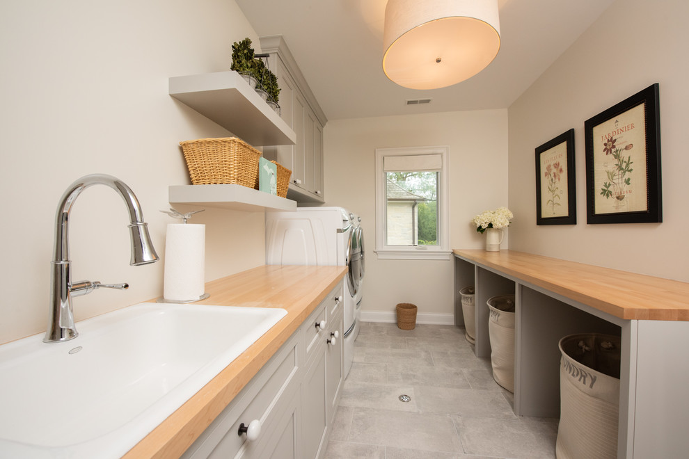 Photo of a medium sized traditional galley utility room in Chicago with a belfast sink, shaker cabinets, grey cabinets, wood worktops, beige walls, ceramic flooring, a side by side washer and dryer, grey floors and beige worktops.