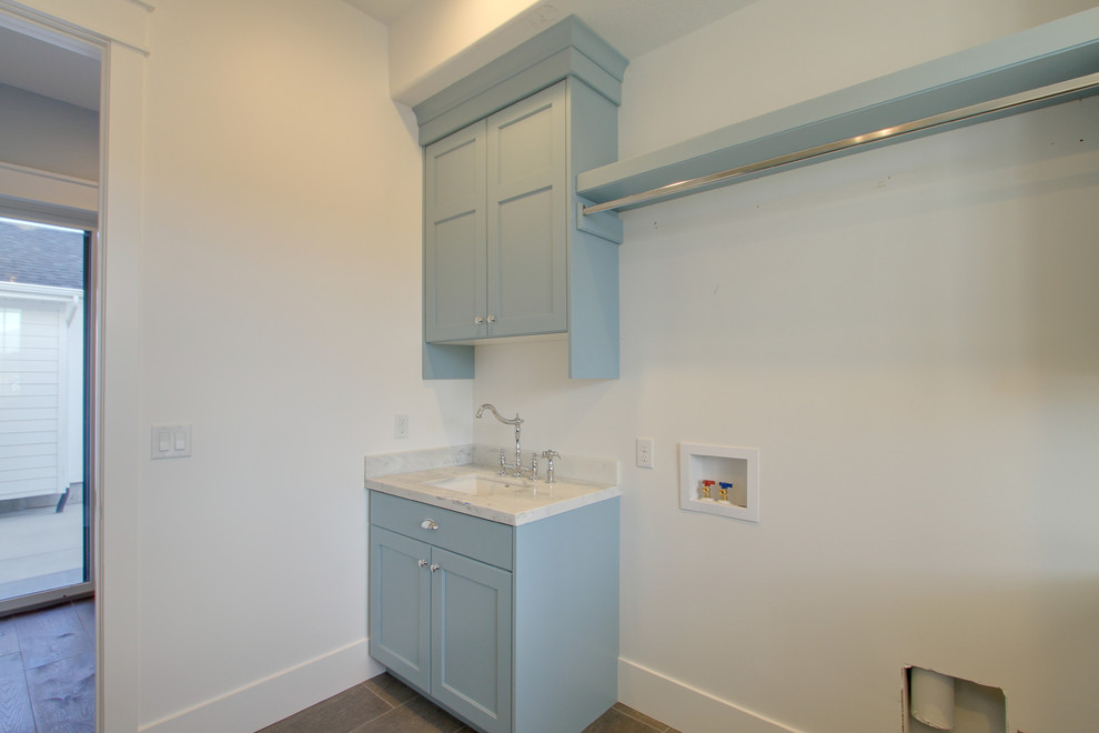 Bild på en mellanstor lantlig u-formad tvättstuga, med en undermonterad diskho, släta luckor, blå skåp, marmorbänkskiva, vita väggar och klinkergolv i porslin