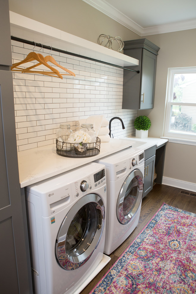 Photo of a medium sized traditional u-shaped utility room in Chicago with a belfast sink, shaker cabinets, engineered stone countertops, white splashback, metro tiled splashback, medium hardwood flooring, brown floors, white worktops, grey cabinets, beige walls and a side by side washer and dryer.