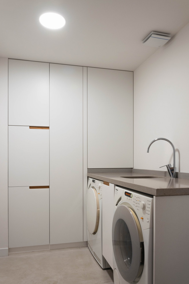 Imagen de cuarto de lavado lineal minimalista de tamaño medio con fregadero bajoencimera, puertas de armario blancas, paredes blancas y suelo gris