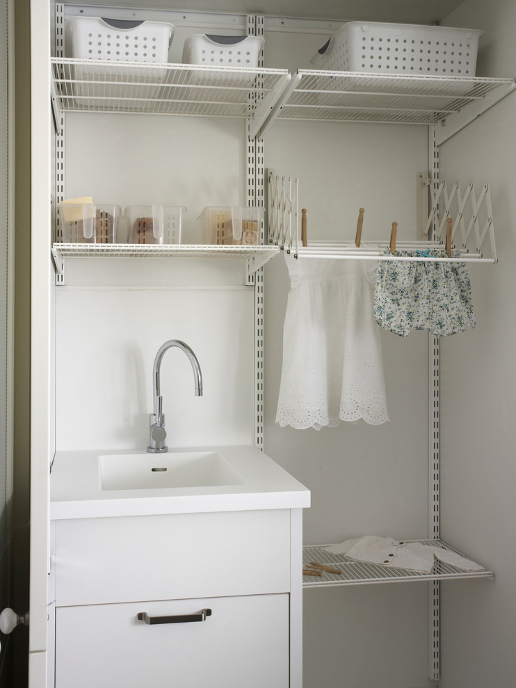 Inredning av en modern liten linjär tvättstuga enbart för tvätt, med vita skåp, vita väggar, ljust trägolv, en tvättmaskin och torktumlare bredvid varandra och en integrerad diskho