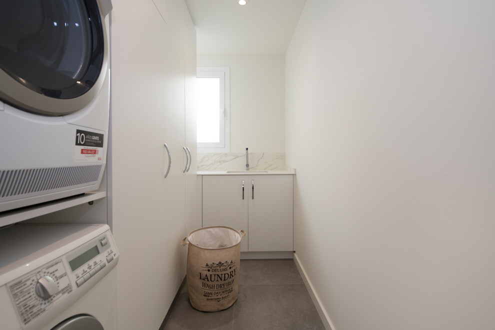 他の地域にある高級な広い北欧スタイルのおしゃれな洗濯室 (L型、シングルシンク、フラットパネル扉のキャビネット、白いキャビネット、クオーツストーンカウンター、白いキッチンパネル、クオーツストーンのキッチンパネル、白い壁、磁器タイルの床、上下配置の洗濯機・乾燥機、グレーの床、白いキッチンカウンター) の写真