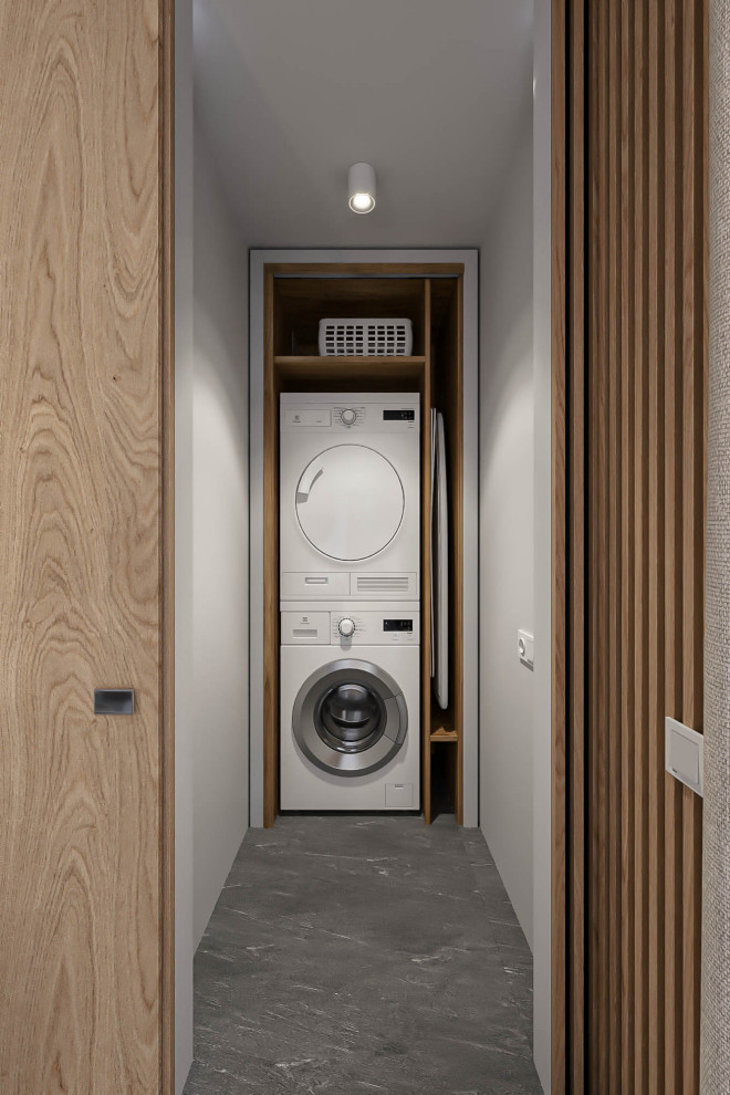 Modelo de armario lavadero lineal moderno pequeño con armarios abiertos, puertas de armario de madera oscura, paredes blancas, suelo de baldosas de porcelana, lavadora y secadora apiladas y suelo gris