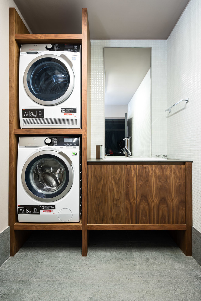 Bild på en liten funkis svarta linjär svart tvättstuga enbart för tvätt, med en integrerad diskho, öppna hyllor, skåp i mellenmörkt trä, bänkskiva i kvarts, vita väggar, skiffergolv, en tvättpelare och grått golv